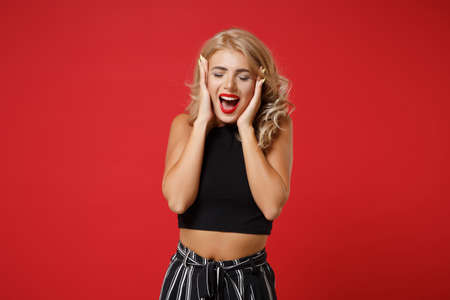 Overjoyed young woman girl in black clothes posing isolated on red background studio portrait. People sincere emotions lifestyle concept. Mock up copy space. Keeping eyes closed, put hands on cheeks.の写真素材