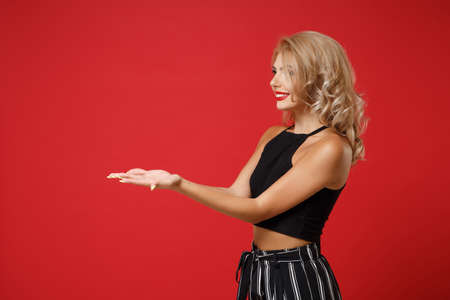Side view of smiling pretty young woman girl in black clothes posing isolated on bright red wall background, studio portrait. People lifestyle concept. Mock up copy space. Holding something in hands.の写真素材
