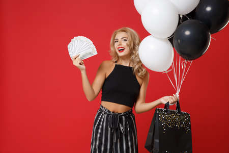 Smiling young woman girl in black clothes posing isolated on red wall background. Shopping discount sale concept. Mock up copy space. Holding air balloons package bag with purchases fan of cash money.の写真素材