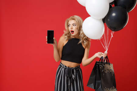 Shocked woman in black clothes posing isolated on red background. Shopping discount sale concept. Mock up copy space. Holding air balloons, package bag with purchases, mobile phone with blank screen.の写真素材