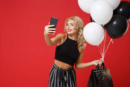 Smiling woman in black clothes posing isolated on red background. Shopping discount sale concept. Mock up copy space. Hold air balloons, package bag with purchases, doing selfie shot on mobile phone.の写真素材