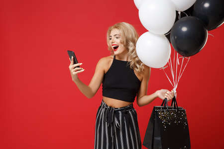 Cheerful young woman girl in black clothes posing isolated on bright red background. Shopping discount sale concept. Mock up copy space. Holding air balloons, package bag with purchases, mobile phone.の写真素材