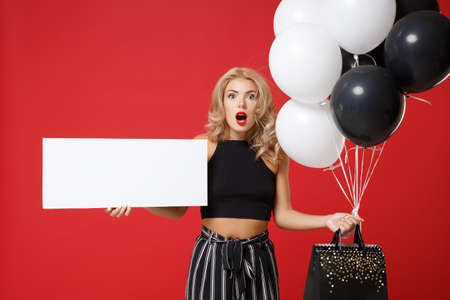 Shocked woman girl in black clothes posing isolated on red background. Shopping discount sale concept. Mock up copy space. Hold air balloons package bag with purchases blank board with place for text.の写真素材