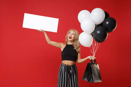 Funny woman girl in black clothes posing isolated on red background. Shopping discount sale concept. Mock up copy space. Hold air balloons package bag with purchases blank board with place for text.の写真素材