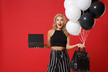 Cheerful young woman girl posing isolated on red background. Valentines Day Womens Day, birthday concept. Mock up copy space. Hold air balloons package bag with purchases black and gold present box.の写真素材