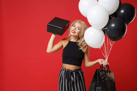 Smiling young woman girl posing isolated on red background. Valentines Day, Womens Day, birthday concept. Mock up copy space. Hold air balloons package bag with purchases black and gold present box.の写真素材