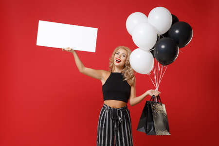 Smiling woman girl in black clothes posing isolated on red background. Shopping discount sale concept. Mock up copy space. Hold air balloons package bag with purchases blank board with place for text.の写真素材