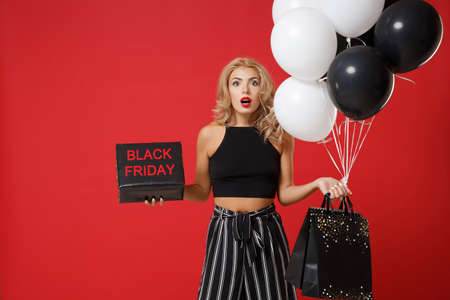 Shocked young woman girl posing isolated on red background. Valentines Day Womens Day, birthday concept. Mock up copy space. Hold air balloons package bag with purchases black and gold present box.の写真素材