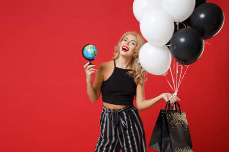 Laughing young woman girl in black clothes posing isolated on red background in studio. Shopping discount sale concept. Mock up copy space. Holding air balloons package bag with purchases world globe.の写真素材