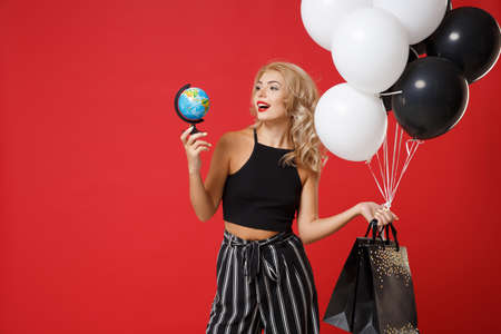 Smiling young woman girl in black clothes posing isolated on red background in studio. Shopping discount sale concept. Mock up copy space. Holding air balloons package bag with purchases, world globe.の写真素材