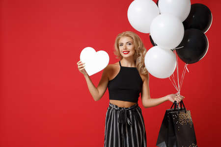 Smiling young woman in black clothes posing isolated on red background. Shopping discount sale concept. Mock up copy space. Holding air balloons package bag with purchases, white blank empty heart.の写真素材