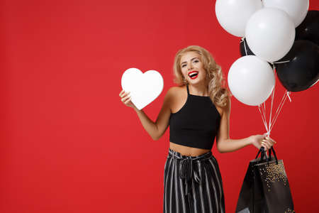 Laughing young woman in black clothes posing isolated on red background. Shopping discount sale concept. Mock up copy space. Holding air balloons package bag with purchases, white blank empty heart.の写真素材