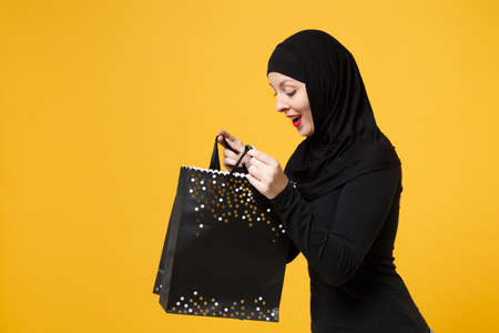Arabian muslim woman in black hijab hold package bag, purchases after shopping isolated on yellow...