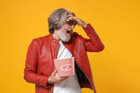 Displeased scared elderly gray-haired mustache bearded man in leather jacket, 3d glasses isolated on yellow background. People in cinema concept. Watch movie film hold popcorn covering eyes with hand.の写真素材