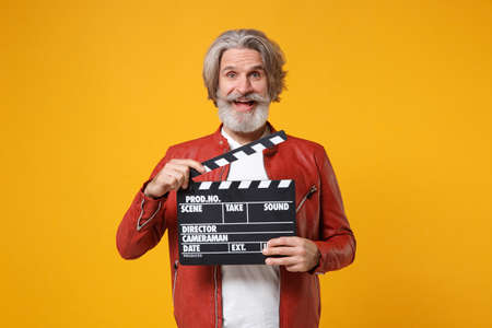 Excited elderly gray-haired mustache bearded man in red leather jacket isolated on yellow orange background. People lifestyle concept. Mock up copy space. Hold classic black film making clapperboard.の写真素材