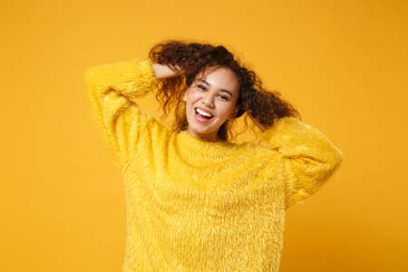 Cheerful young african american girl in fur sweater posing isolated on yellow orange background studio portrait. People sincere emotions lifestyle concept. Mock up copy space. Having fun holding hair.の写真素材