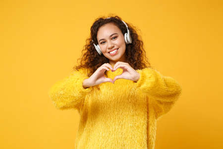Smiling young african american girl in sweater posing isolated on yellow orange background. People lifestyle concept. Mock up copy space. Listen music with headphones, showing shape heart with hands.の写真素材