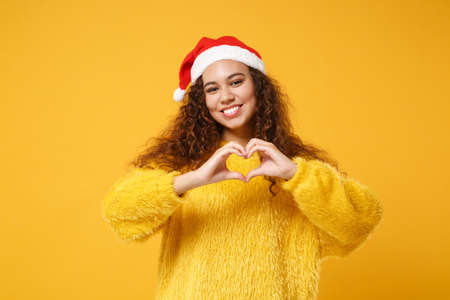 Smiling young african american Santa girl in sweater, Christmas hat isolated on yellow background. Happy New Year 2020 celebration holiday concept. Mock up copy space. Showing shape heart with hands.の写真素材