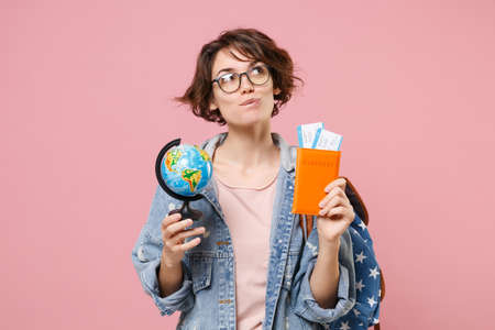 Pensive young woman student in denim clothes eyeglasses backpack isolated on pastel pink background. Education in high school university college concept. Hold passport tickets world globe looking up.の写真素材