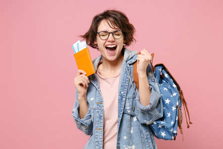Joyful young woman student in denim clothes glasses backpack isolated on pink background. Education in high school university college concept. Hold passport boarding pass tickets doing winner gesture.の写真素材
