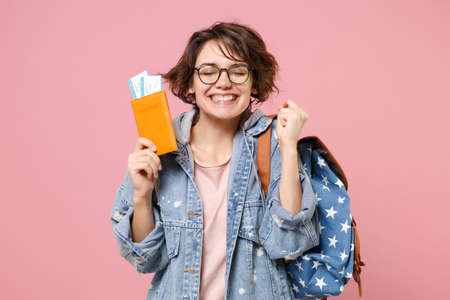 Smiling young woman student in denim clothes glasses backpack isolated on pink background. Education in high school university college concept. Holding passport tickets clenching fist like winner.の写真素材