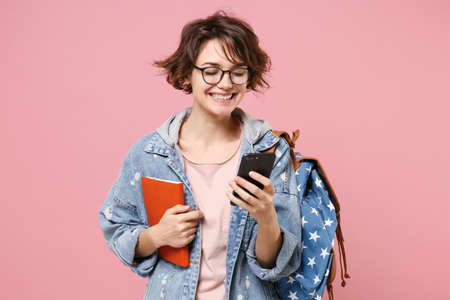 Smiling young woman student in denim clothes glasses backpack isolated on pastel pink background. Education in high school university college concept. Hold books using mobile phone typing sms message.の写真素材