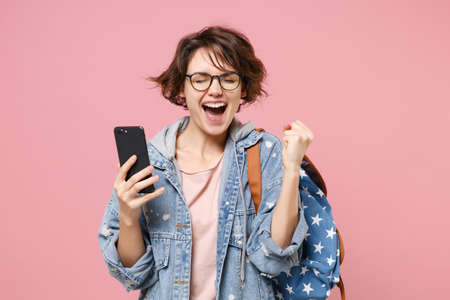 Joyful woman student in denim clothes glasses backpack isolated on pastel pink background. Education in school university college concept. Using mobile phone typing sms message doing winner gesture.の写真素材