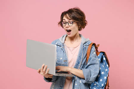Excited young woman student in denim clothes glasses backpack isolated on pastel pink background. Education in high school university college concept. Mock up copy space. Work on laptop pc computer.の写真素材