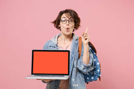 Amazed girl student in denim clothes glasses backpack isolated on pink background. Education in high school university college concept. Hold laptop computer with blank screen pointing index finger up.の写真素材