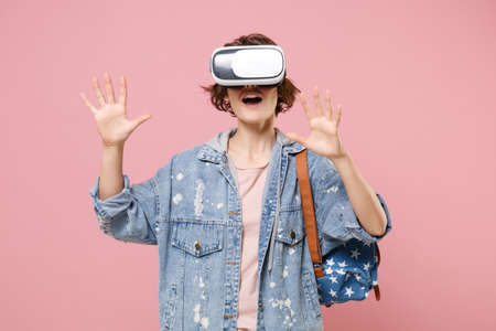 Young woman student in denim clothes backpack isolated on pastel pink background. Education in high school university college concept. Watching in headset, touch something like push click on button.の写真素材