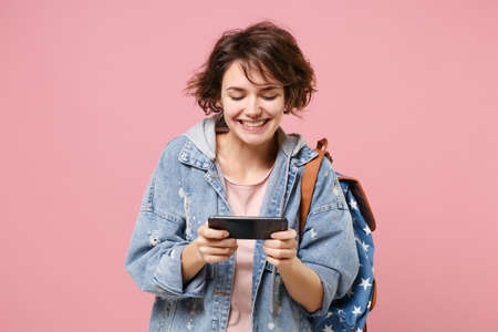 Smiling young girl student in denim clothes backpack isolated on pastel pink background in studio. Education in high school university college concept. Mock up copy space. Play game with mobile phone.の写真素材