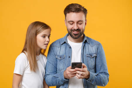 Shocked bearded man in casual clothes with child baby girl. Father little kid daughter isolated on yellow background. Love family day parenthood childhood concept. Use mobile phone typing sms message.の写真素材