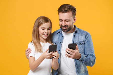 Smiling man in casual clothes with child baby girl. Father little kid daughter isolated on yellow background. Love family day parenthood childhood concept. Use mobile phone typing sms message hugging.の写真素材