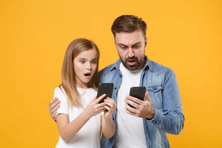 Shocked man in casual clothes with child baby girl. Father little kid daughter isolated on yellow background. Love family day parenthood childhood concept. Use mobile phone typing sms message hugging.の写真素材
