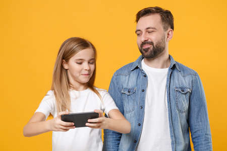 Funny bearded man in casual clothes have fun with child baby girl. Father little kid daughter isolated on yellow background. Love family day parenthood childhood concept. Play game with mobile phone.の写真素材