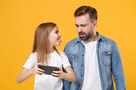 Shocked bearded man in casual clothes have fun with child baby girl. Father little kid daughter isolated on yellow background. Love family day parenthood childhood concept Play game with mobile phone.の写真素材
