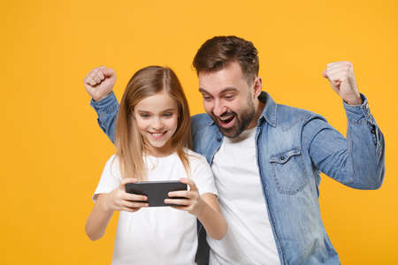Joyful bearded man with cute child baby girl. Father little kid daughter isolated on yellow background. Love family day parenthood childhood concept. Play game with mobile phone, doing winner gesture.の写真素材
