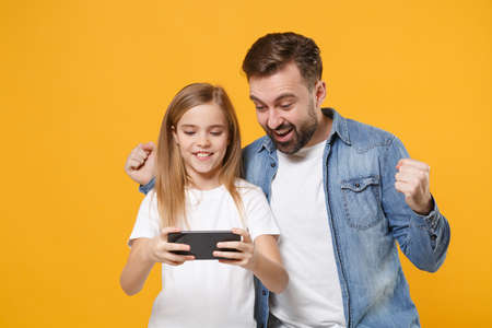 Joyful bearded man with cute child baby girl. Father little kid daughter isolated on yellow background. Love family day parenthood childhood concept. Play game with mobile phone, doing winner gesture.の写真素材