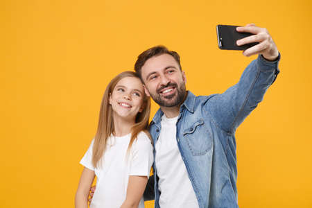 Smiling bearded man in casual clothes with child baby girl. Father little kid daughter isolated on yellow background. Love family day parenthood childhood concept. Doing selfie shot on mobile phone.の写真素材