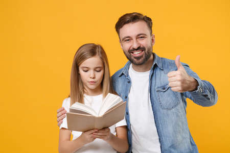 Smiling bearded man in casual clothes with cute child baby girl. Father little kid daughter isolated on yellow background. Love family day parenthood childhood concept. Reading book, showing thumb up.の写真素材