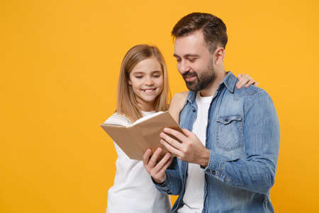 Pretty bearded man in casual clothes have fun with cute child baby girl. Father little kid daughter isolated on yellow background in studio. Love family day parenthood childhood concept. Reading book.の写真素材