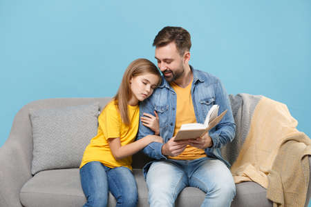 Smiling bearded man in casual clothes with cute child baby girl. Father little daughter isolated on pastel blue background. Love family parenthood childhood concept. Sit on couch reading book hugging.の写真素材