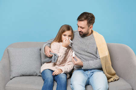 Man in knitted sweater with sick child baby girl. Father little daughter isolated on blue background. Love family parenthood childhood concept. Sit on couch, having runny nose, blowing nose to napkin.の写真素材