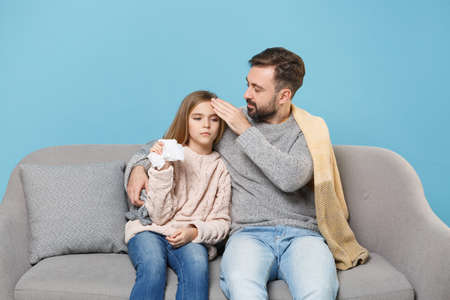 Bearded man in knitted sweater with sick tired child baby girl. Father little daughter isolated on blue background. Love family parenthood childhood concept. Sitting on couch, holding paper napkin.の写真素材