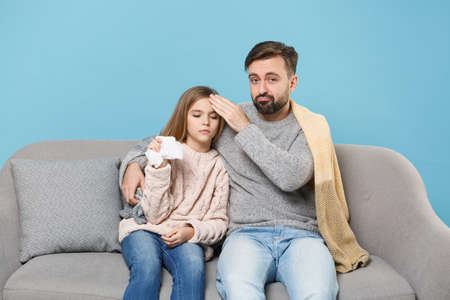 Bearded man in knitted sweater with sick tired child baby girl. Father little daughter isolated on blue background. Love family parenthood childhood concept. Sitting on couch, holding paper napkin.の写真素材