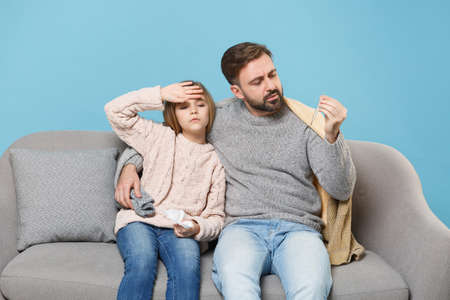 Bearded man in knitted sweater with sick tired child baby girl. Father little daughter isolated on blue background. Love family parenthood childhood concept. Sitting on couch hold napkin, thermometer.の写真素材