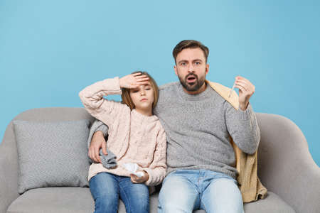 Bearded man in knitted sweater with sick tired child baby girl. Father little daughter isolated on blue background. Love family parenthood childhood concept. Sitting on couch hold napkin, thermometer.の写真素材