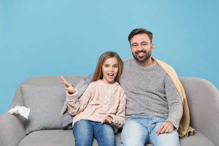 Cheerful bearded man in knitted sweater with child baby girl. Father little daughter isolated on pastel blue background. Love family parenthood childhood concept. Sitting on couch, spreading hands.の写真素材