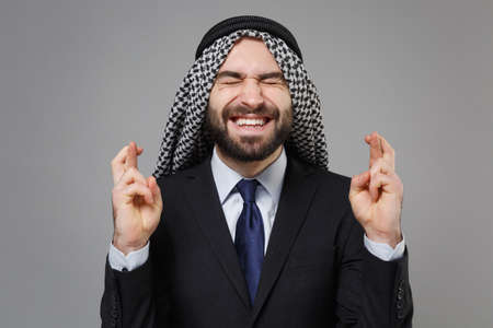 Arabian muslim businessman in keffiyeh kafiya ring igal agal isolated on gray background. Achievement career wealth business. Wait for special moment, keeping fingers crossed, eyes closed, make wish.の写真素材