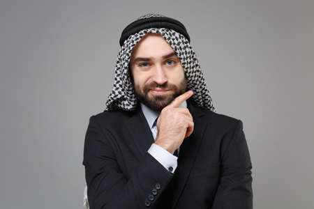 Confused bearded arabian muslim businessman in keffiyeh kafiya ring igal agal classic black suit shirt isolated on gray background. Achievement career wealth business concept Put hand prop up on chin.の写真素材